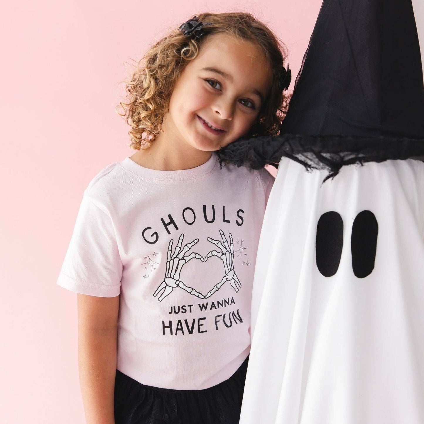 A young girl with curly brown hair smiles next to a large ghost costume prop. She is wearing a light pink graphic t-shirt with black text that reads 'GHOULS JUST WANNA HAVE FUN' and a skeleton hand graphic making a heart shape.