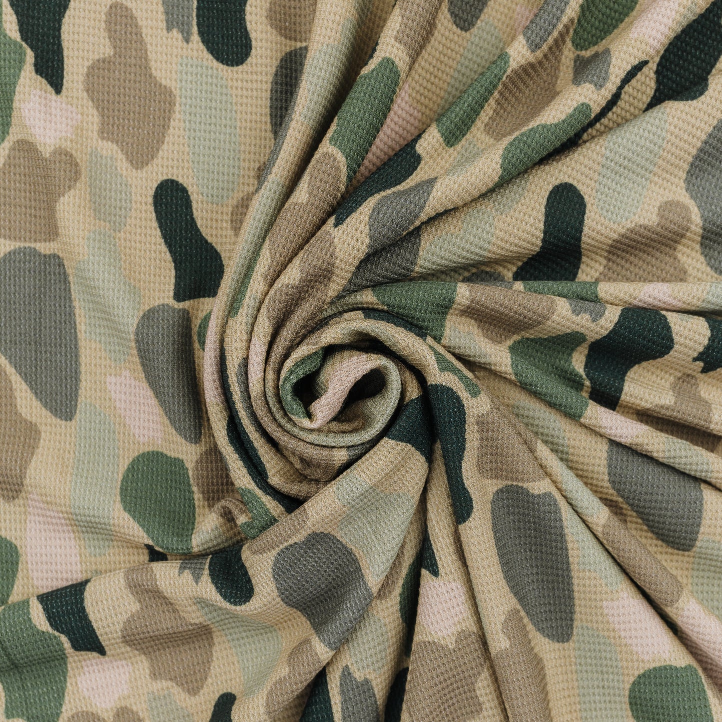 A close-up of camouflage-patterned blanket arranged in a spiral drape, showing a textured waffle-weave surface with shades of beige, green, and gray.