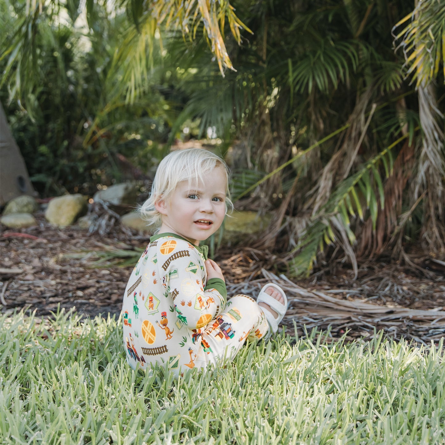 Baby wearing All Aboard the Dinosaur Train™ bamboo pajamas sitting on grass outdoors