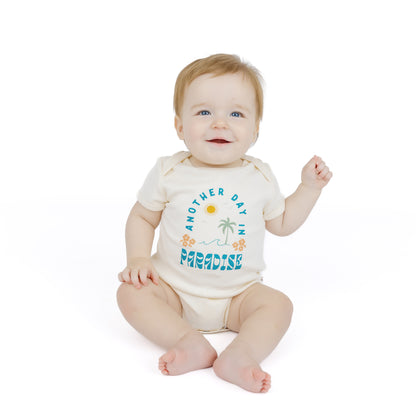 Baby sitting on a white background wearing a bodysuit with text and graphics saying Another day in Paradise.