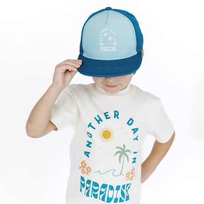 Child wearing a blue and light blue cap and a white t-shirt with a tropical design.