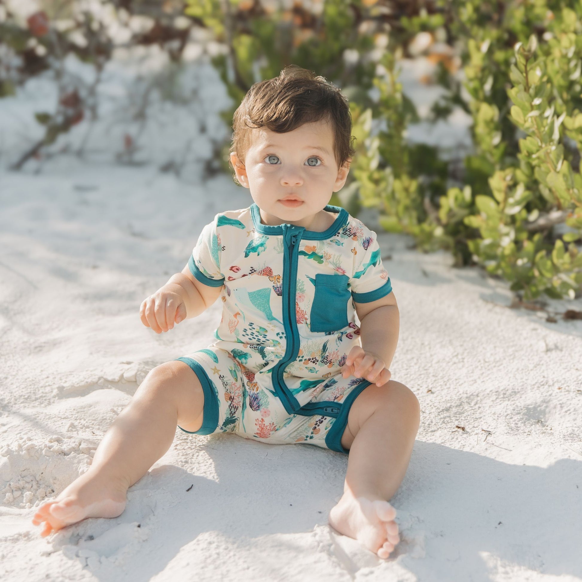Baby girl sitting at the beach sand wearing Emerson and Friends Coral Reef Bamboo Baby Shortie Rompers, featuring a breathable ocean-themed print with sharks, sea turtles, and coral, designed for summer comfort and easy zip-up wear.