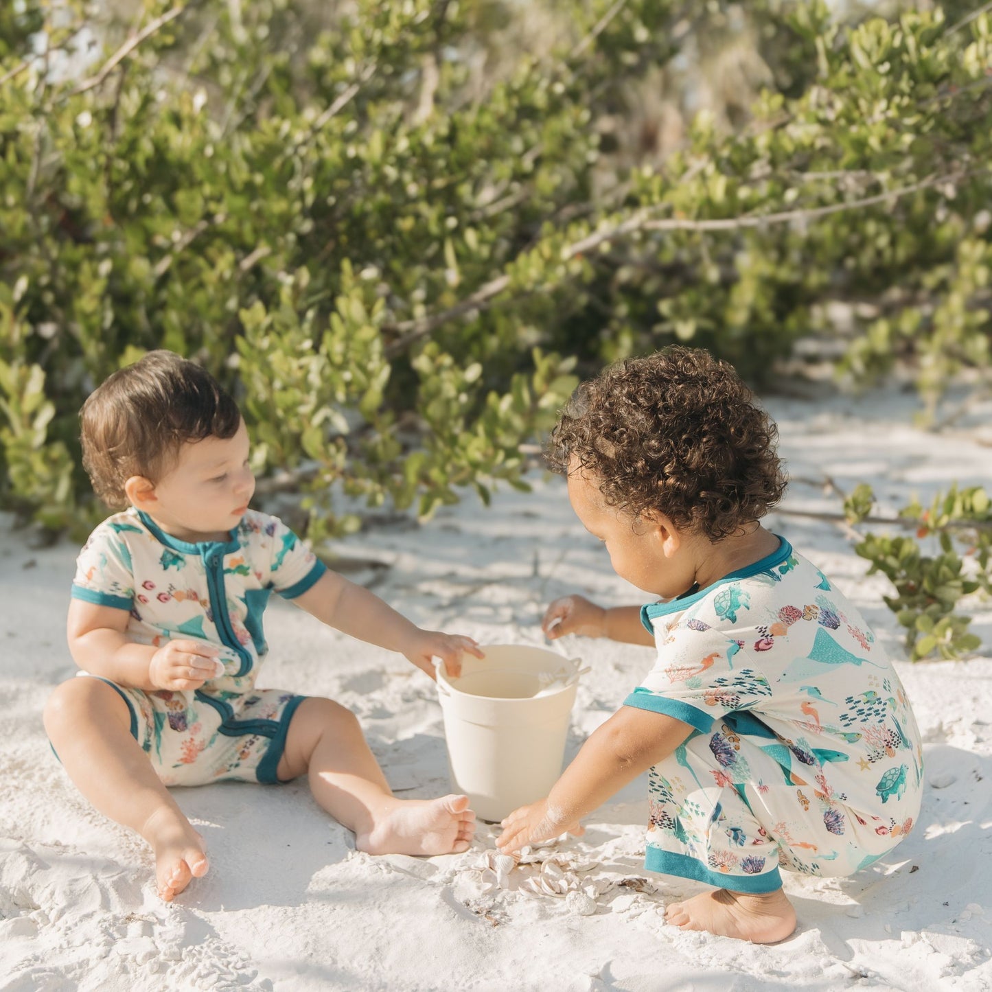Two babies playing in beach sand wearing Emerson and Friends Coral Reef Bamboo Baby Shortie Rompers, featuring a breathable ocean-themed print with sharks, sea turtles, and coral, designed for summer comfort and easy zip-up wear.