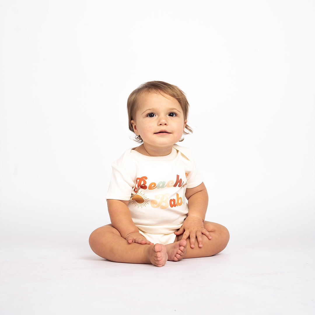 a baby sitting on the floor in the "beach babe" cotton bodysuit. the word 'beach babe' is spelt out in a groovy font with a sun, starfish, and shell surrounding it. 