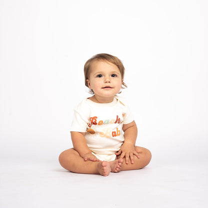 a baby sitting on the floor in the "beach babe" cotton bodysuit. the word 'beach babe' is spelt out in a groovy font with a sun, starfish, and shell surrounding it. 