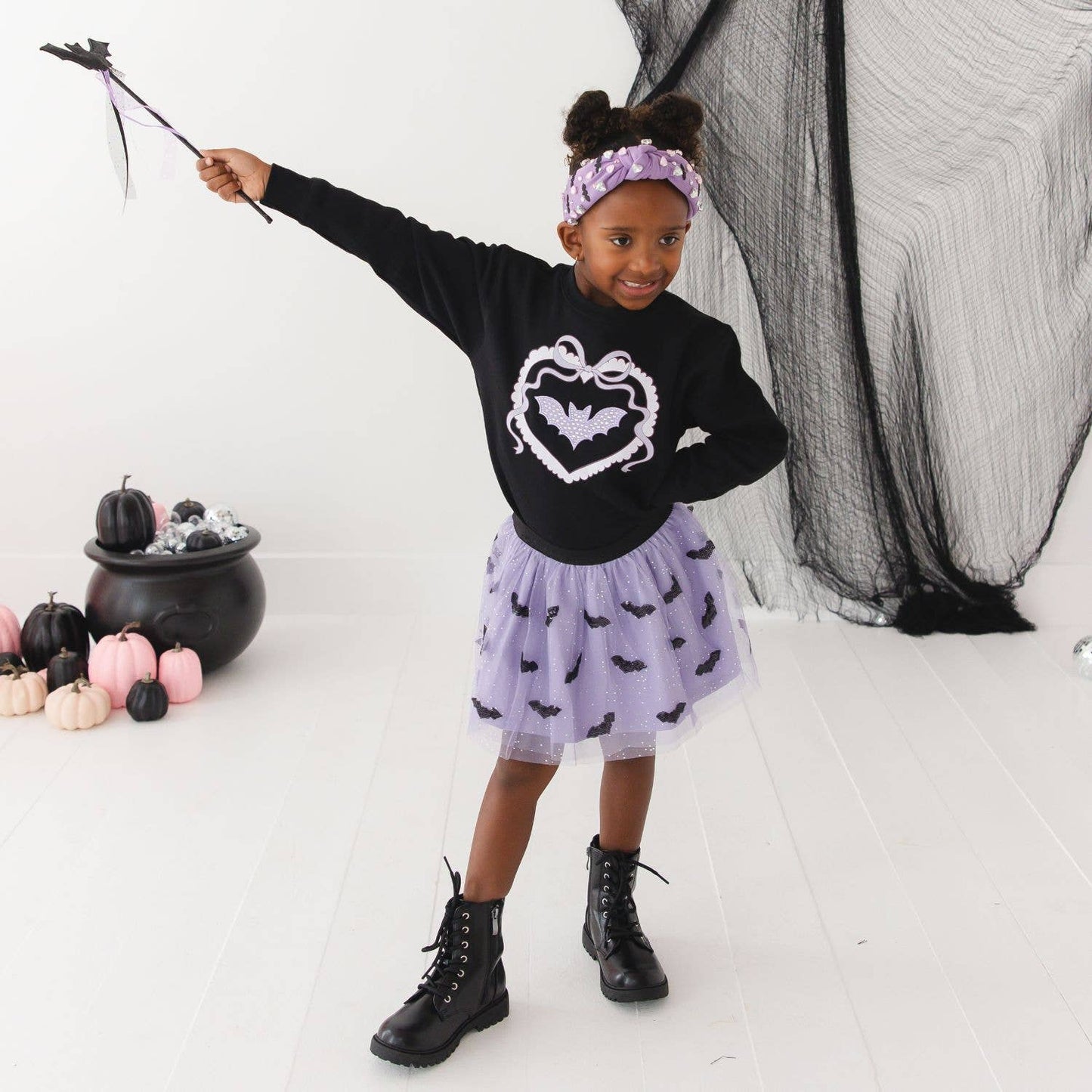 A young girl modeling a black long-sleeve shirt with a white bat-heart graphic and a light purple tutu skirt printed with small black bats. She is holding a witch's wand and wearing a purple patterned headband, standing in a white studio decorated with Halloween props like a cauldron and black netting.