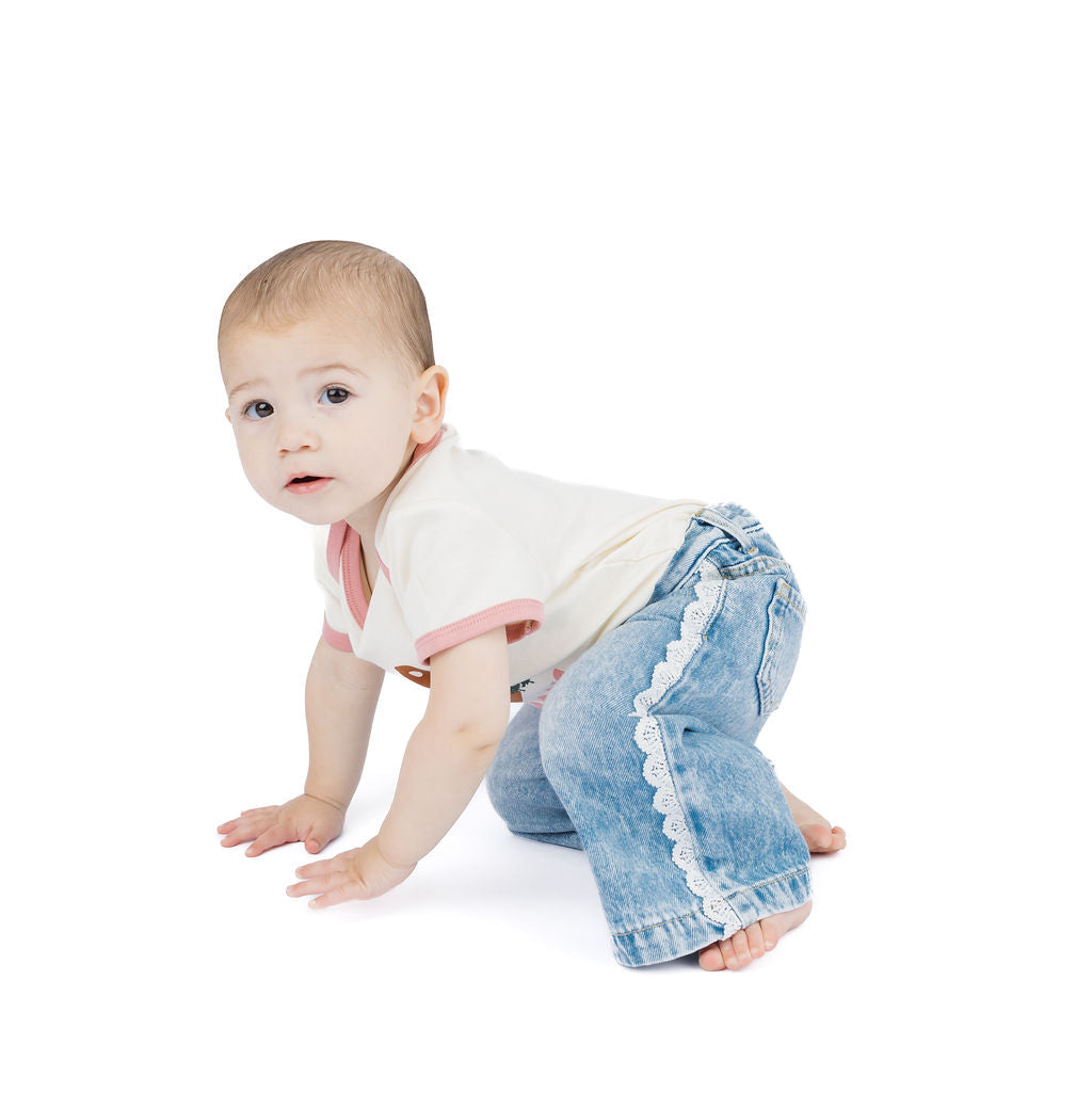 A baby crawling on a white studio background, wearing a cream ringer bodysuit with pink trim and light blue denim jeans with white lace trim down the leg.