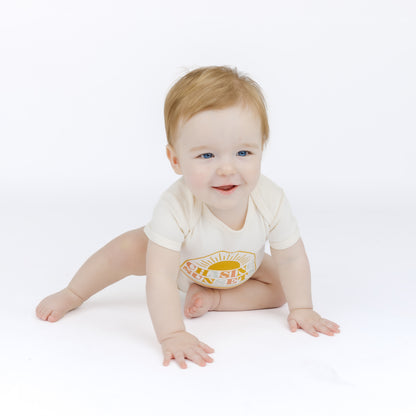 Baby crawling on a white surface wearing a white bodysuit with a colorful design.