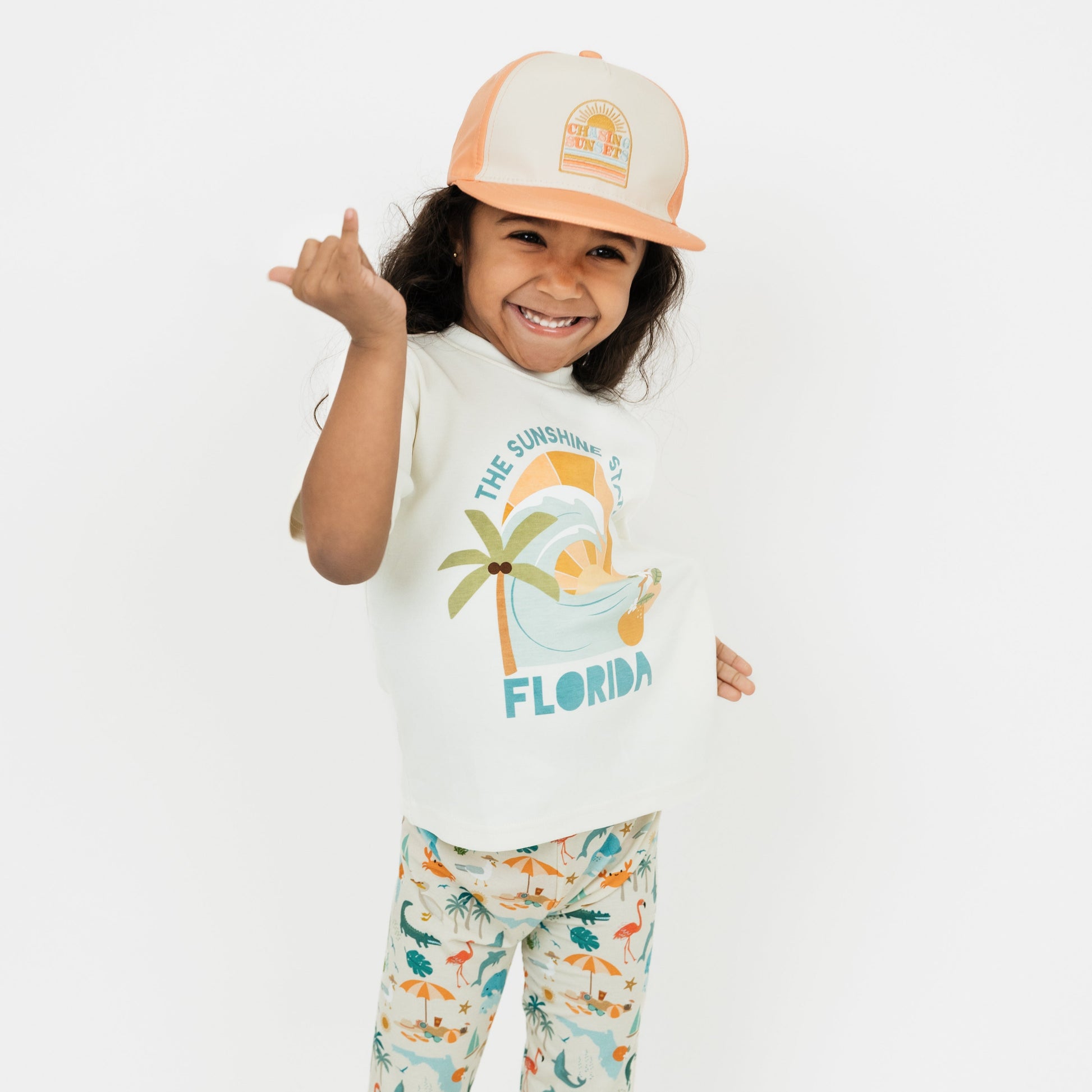 Little girl wearing our Chasing Sunsets Snapback Hat and The Sunshine State Florida Cotton Tee and Explore Florida Bamboo PJ pants holding up a shaka hand gesture.