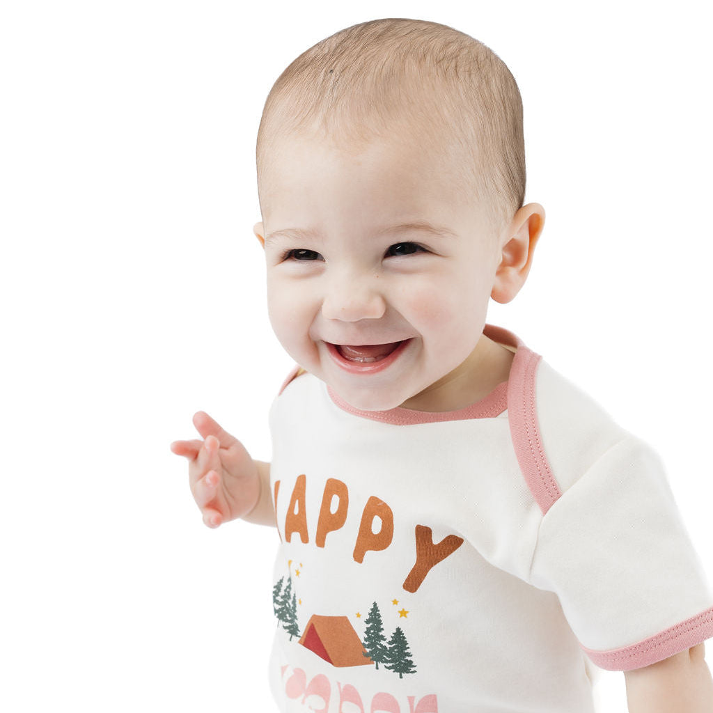 A smiling baby wearing a white bodysuit with pink trim and a 'HAPPY CREEP' graphic, paired with light blue denim jeans, set against a plain white background.