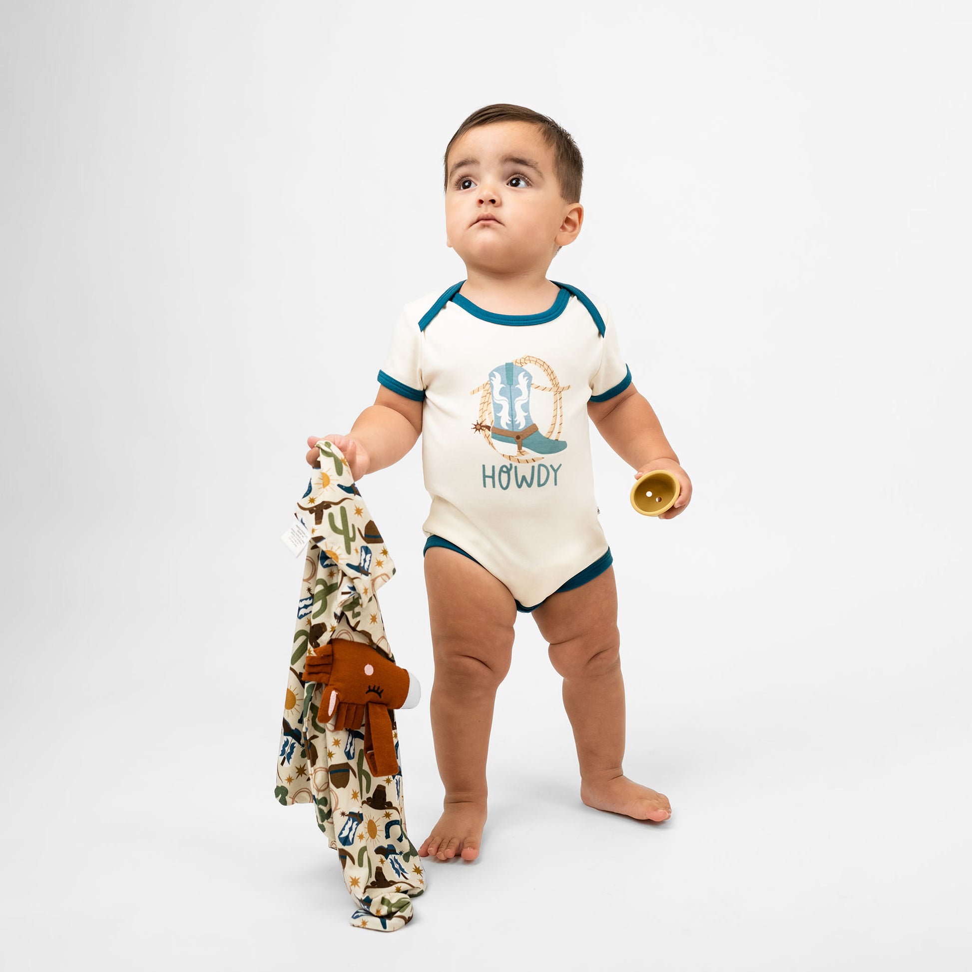 Baby boy wearing our Howdy cotton bodysuit featuring the word "Howdy" in playful, blue letters, perfect for a western outfit while holding our Howdy Lovey