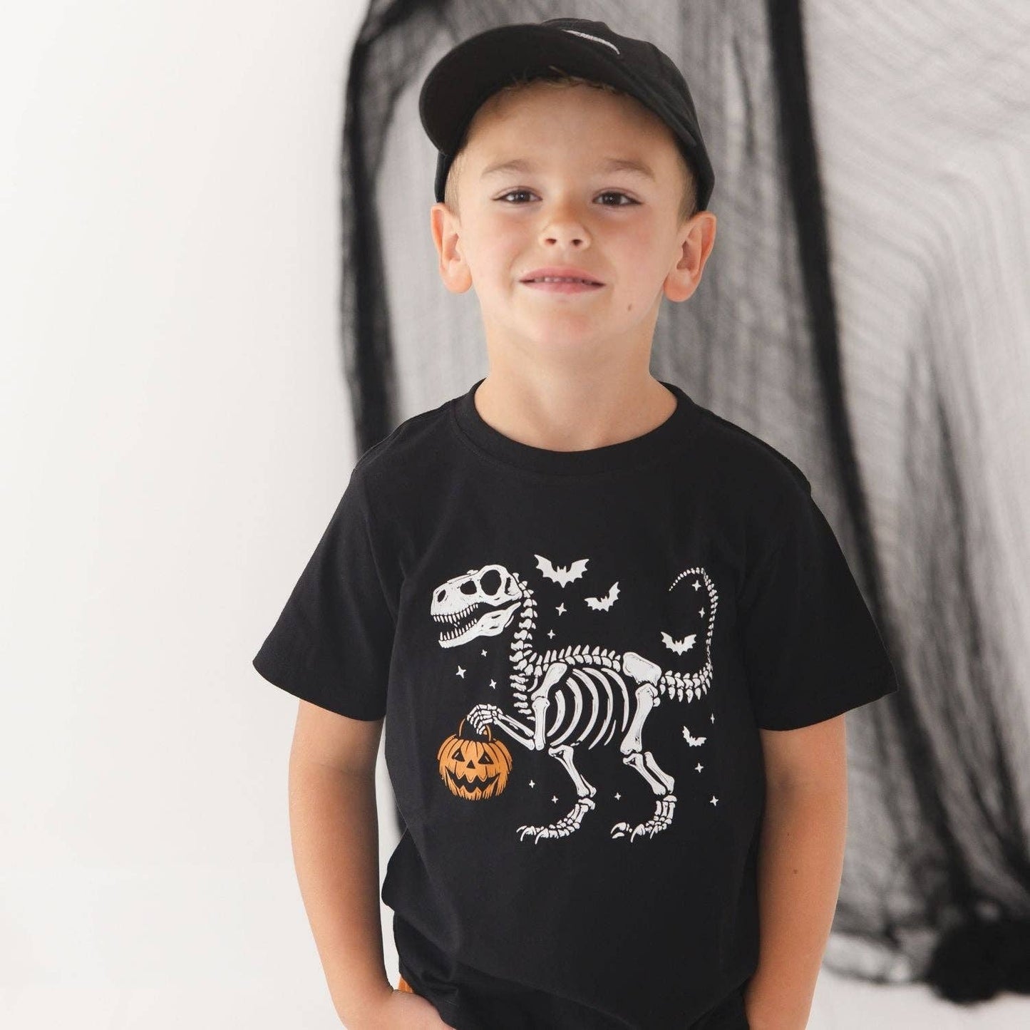 A young boy wearing a black t-shirt with a white dinosaur skeleton and orange pumpkin graphic, paired with a black baseball cap, standing against a white background with a black spiderweb decoration.