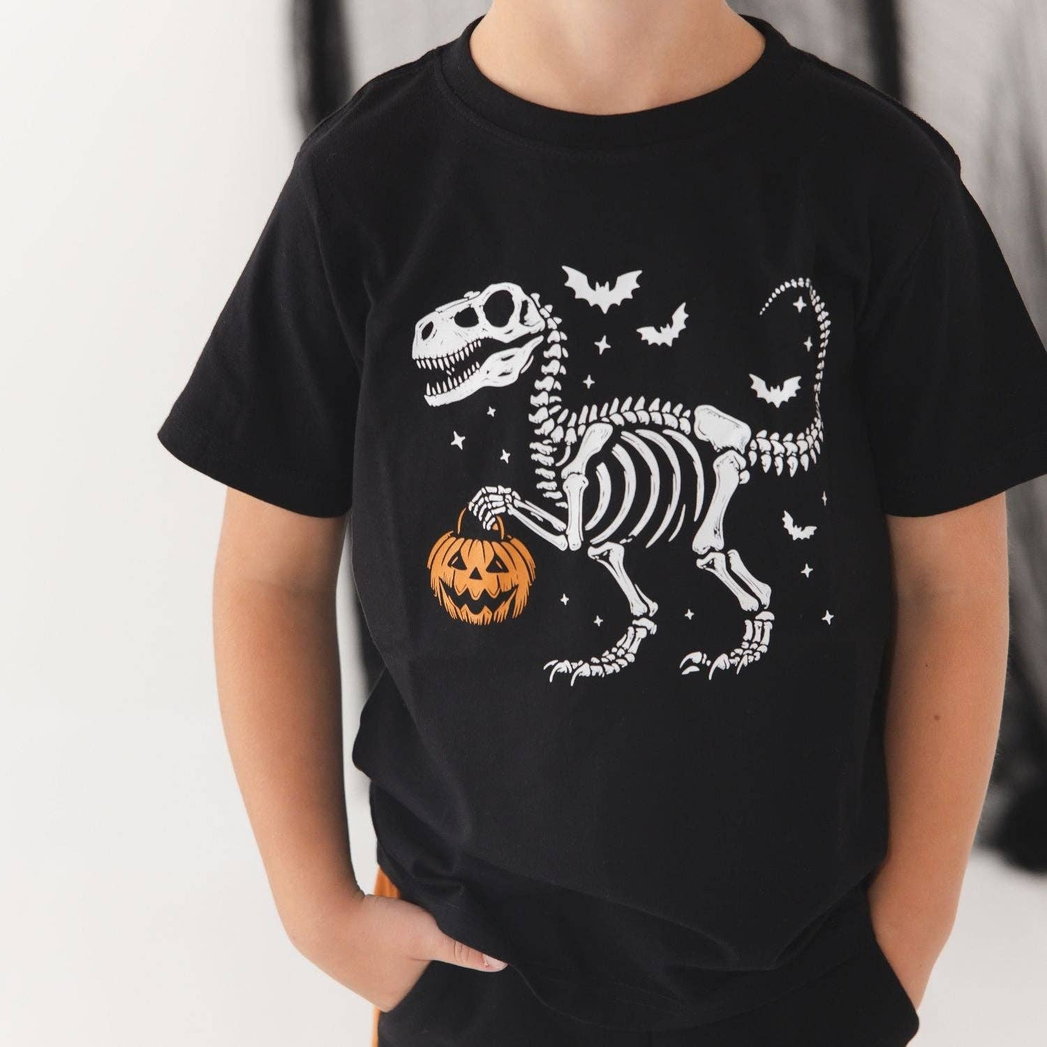 A child wearing a black short-sleeve T-shirt with a white skeleton dinosaur graphic holding an orange jack-o'-lantern, with small white bats and stars around it.