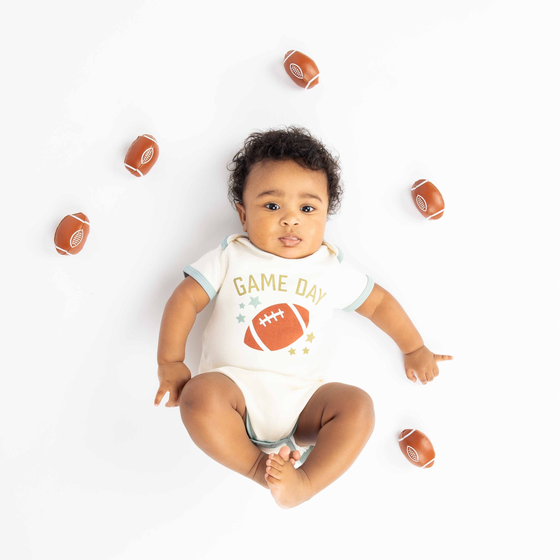 baby laying down with footballs around him. Modeling a blue ringer white bodysuit with a football, stars and the phrase "game Day" on it. 
