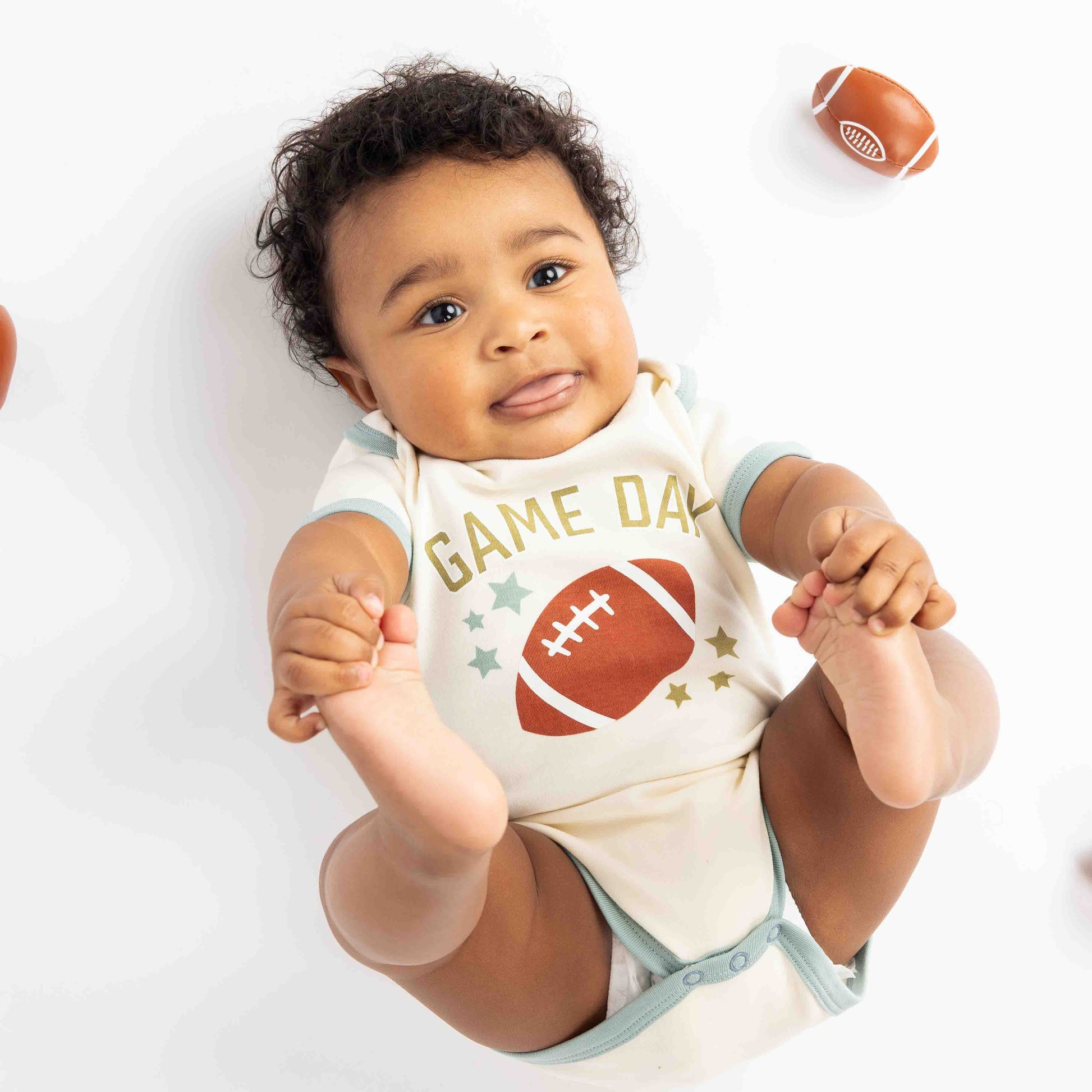 baby laying down with footballs around him. Modeling a blue ringer white bodysuit with a football, stars and the phrase "game Day" on it. 