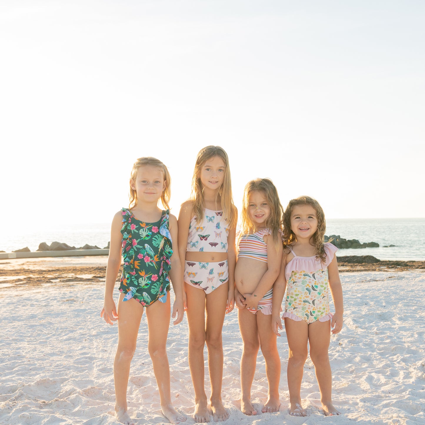 A group of girls on the beach wearing various styles from the Isla Botánica swim collection, including the ruffle one-piece and tankini.