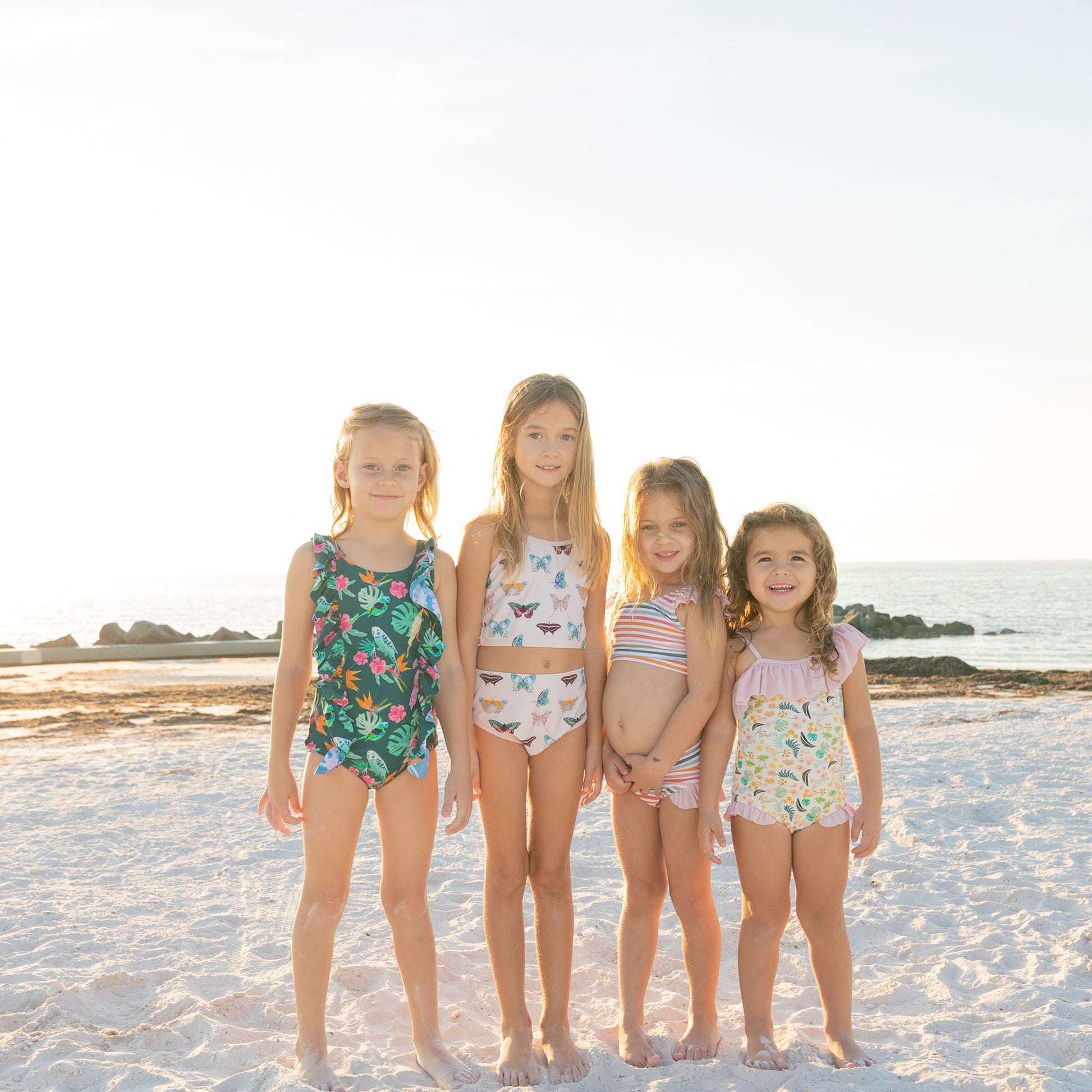A group of girls on the beach wearing various styles from the Isla Botánica swim collection, including the ruffle one-piece and tankini.