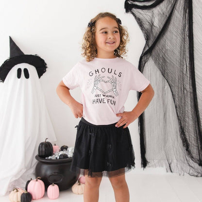 A young girl with curly hair stands in a Halloween-themed room wearing a light pink graphic t-shirt that reads 'Ghouls Just Wanna Have Fun' and a black tulle skirt. She is posed in front of a white ghost decoration, black netting, and pumpkins.
