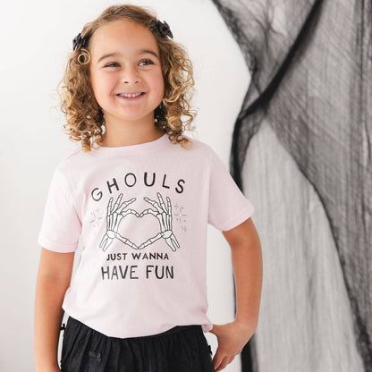 A young girl with curly hair wearing a light pink graphic t-shirt that reads 'GHOULS JUST WANNA HAVE FUN' with skeleton hands forming a heart, posed against a white and black draped background.