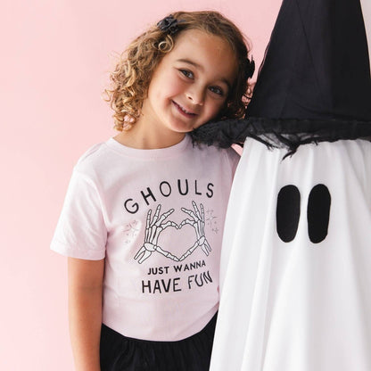 A young girl with curly brown hair smiles next to a large ghost costume prop. She is wearing a light pink graphic t-shirt with black text that reads 'GHOULS JUST WANNA HAVE FUN' and a skeleton hand graphic making a heart shape.