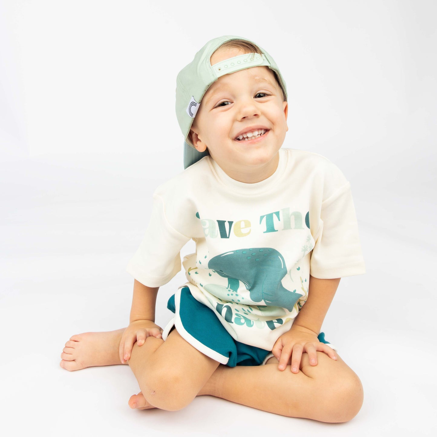 Little boy sitting with the Happiness Comes in Waves hat on backwards while wearing the Emerson and Friends Save the Manatees Cotton Tee with terry cloth shorts. 