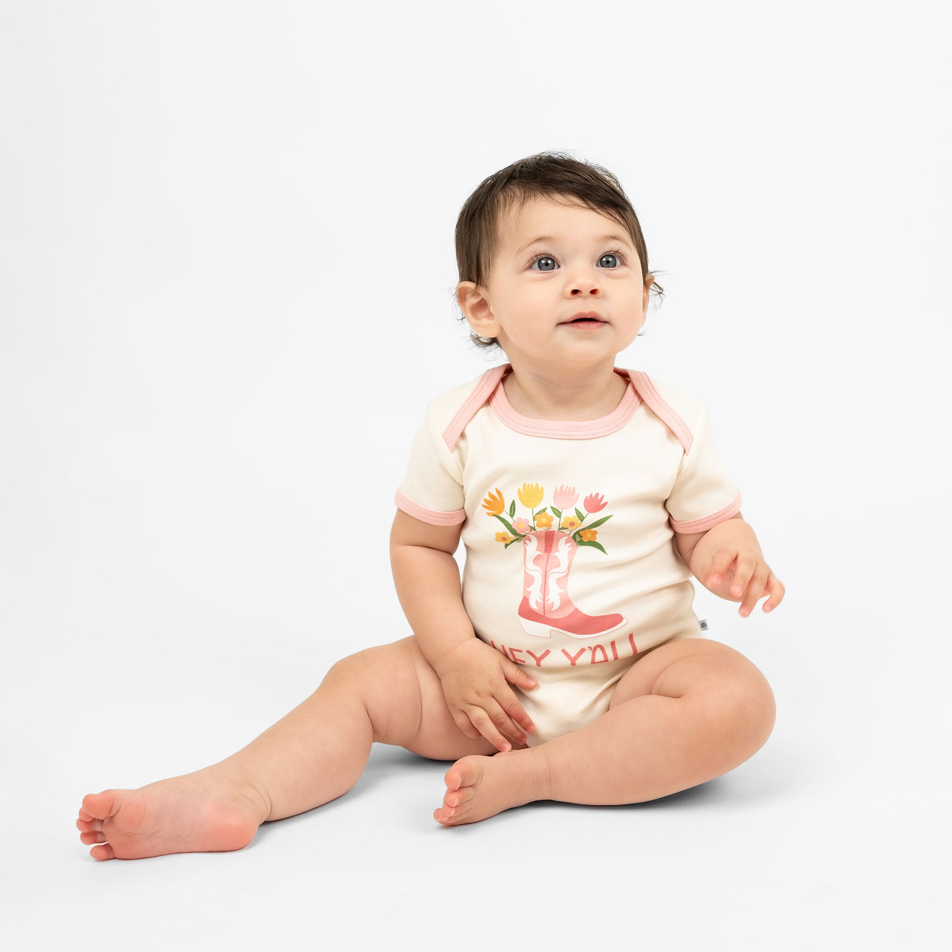 A cute baby wearing our Hey Y'all bodysuit paired with a pink cowgirl boots print and floral accents, capturing the essence of rodeo and western style for a perfect outfit.