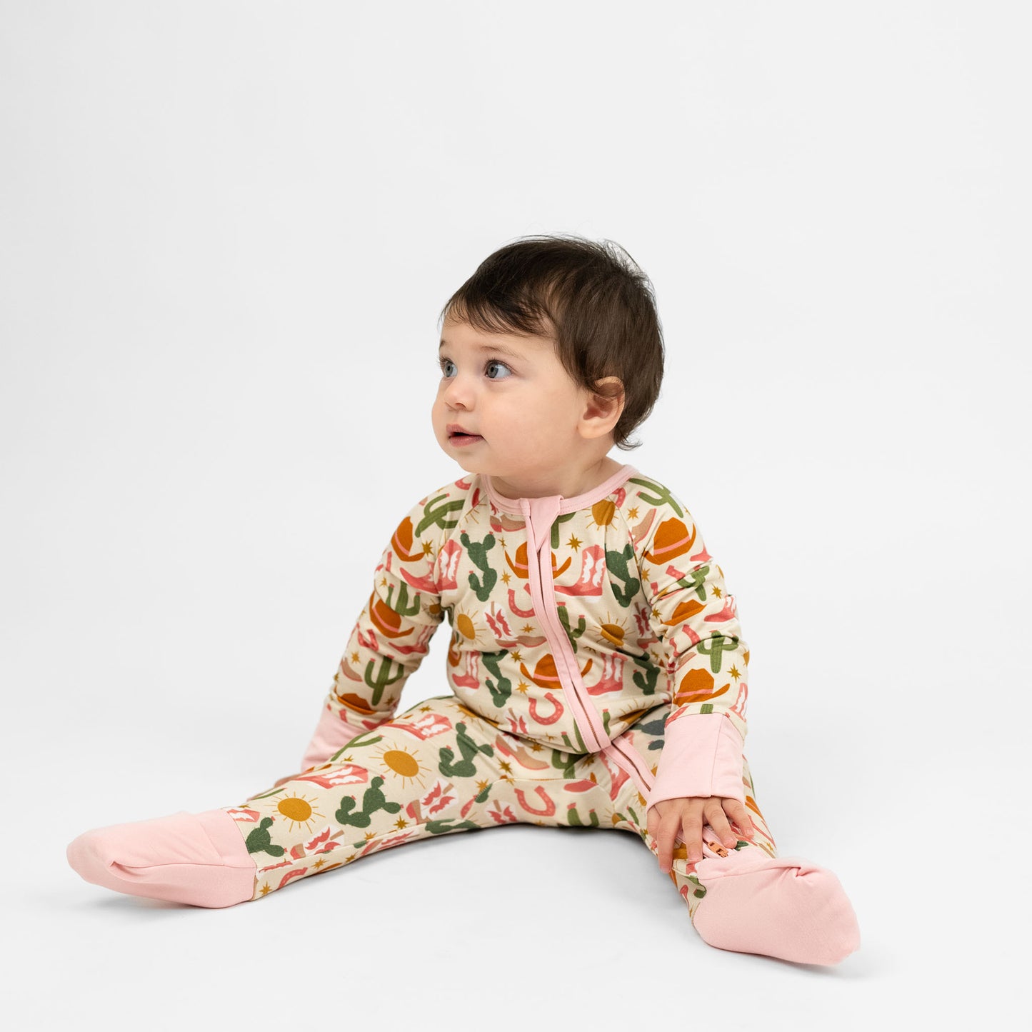 Baby girl wearing our Emerson and Friends Hey Y’all Bamboo Convertible Pajamas. The print shows  green cactus, yellow sun, brown cowgirl hat, pink boots and horseshoes. Perfect for cozy rodeo nights or pajama parties with a cowgirl flair!