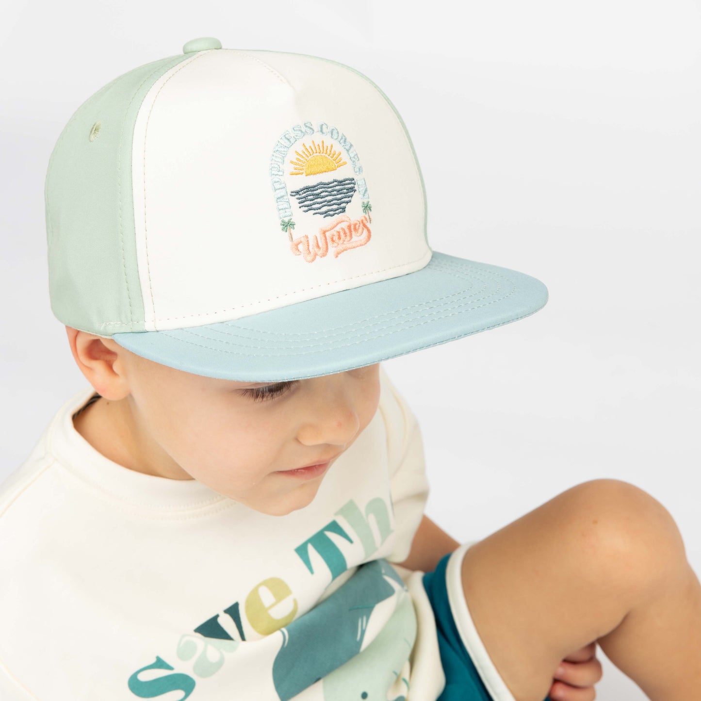 Little boy sitting with the Happiness Comes in Waves hat on backwards while wearing the Emerson and Friends Save the Manatees Cotton Tee with terry cloth shorts. 