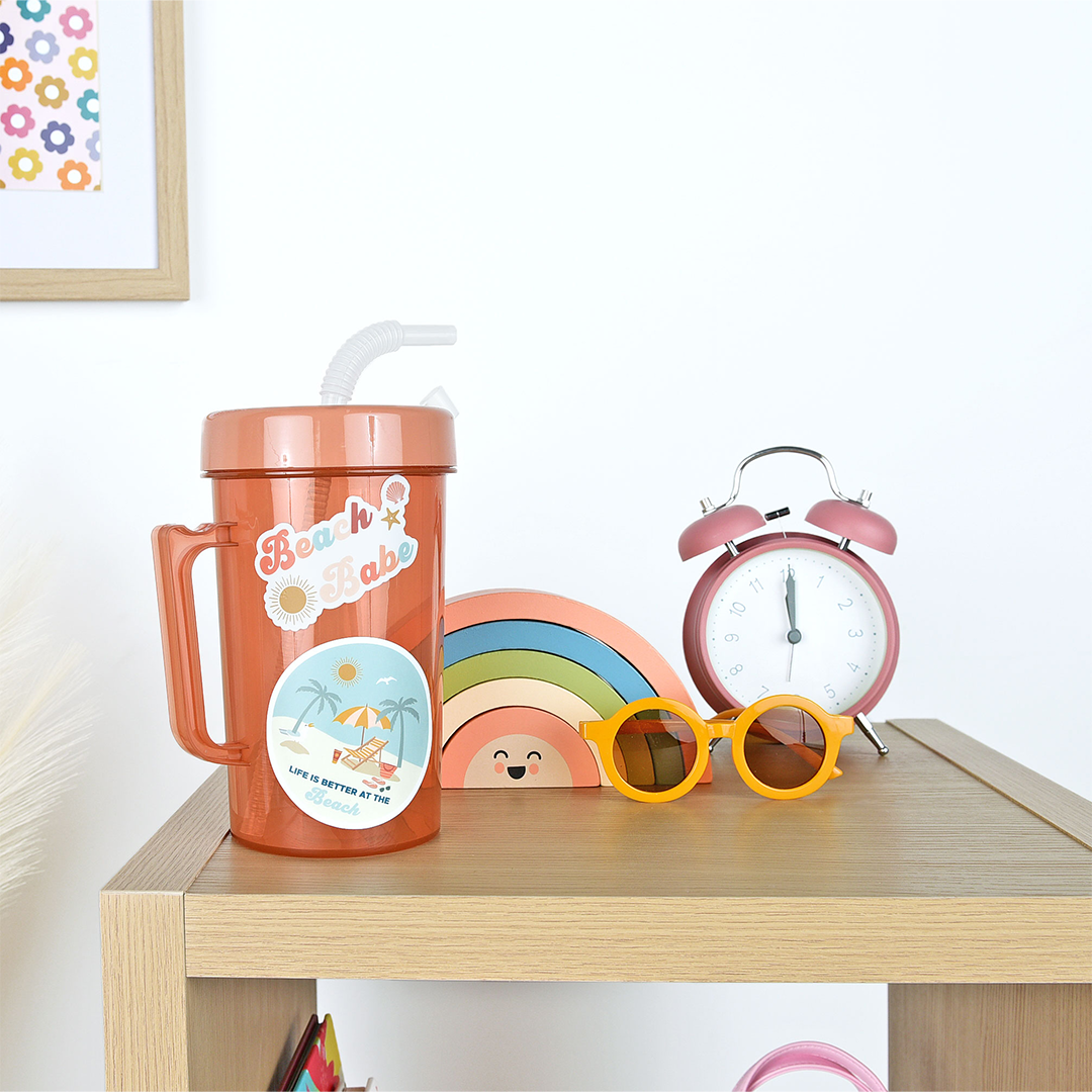 a water bottle is on a table. the water bottle has the "beach babe" sticker on it and the "life is better at the beach" sticker. 