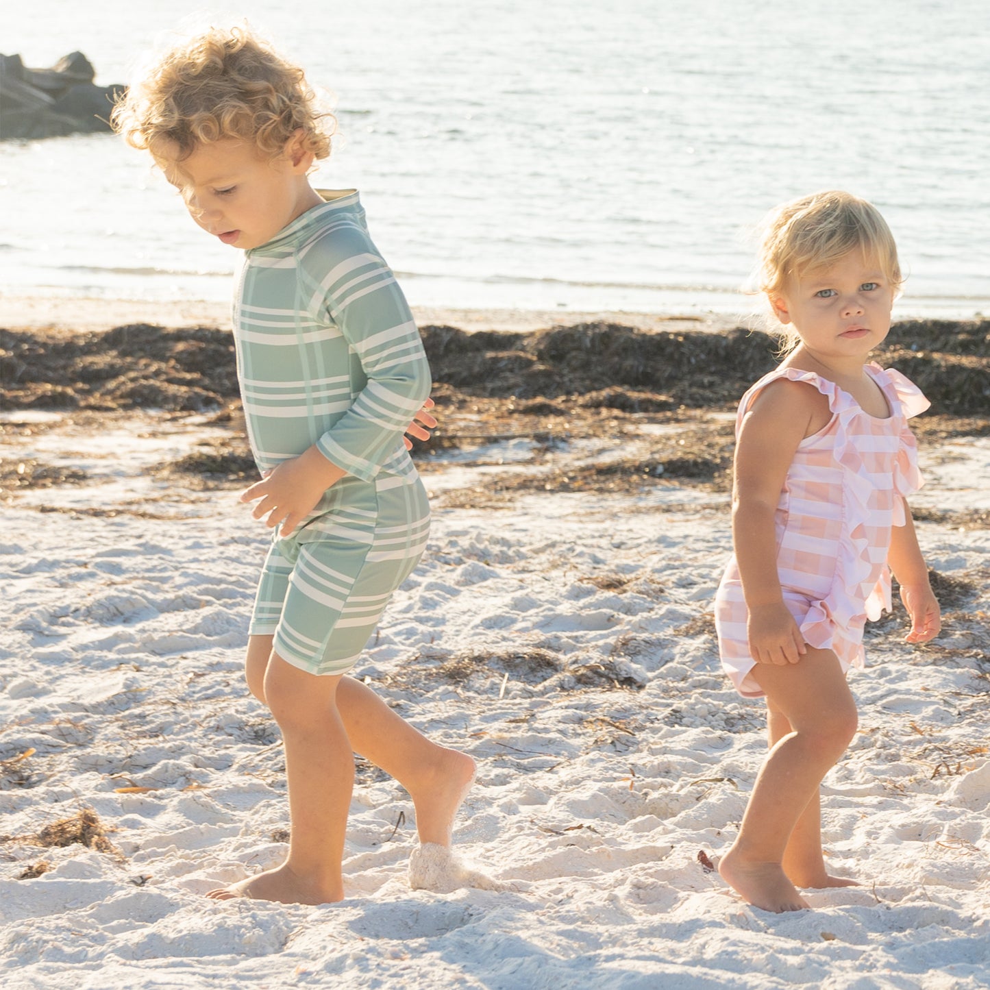 Two children in swimsuits standing on a sandy beach with water in the background