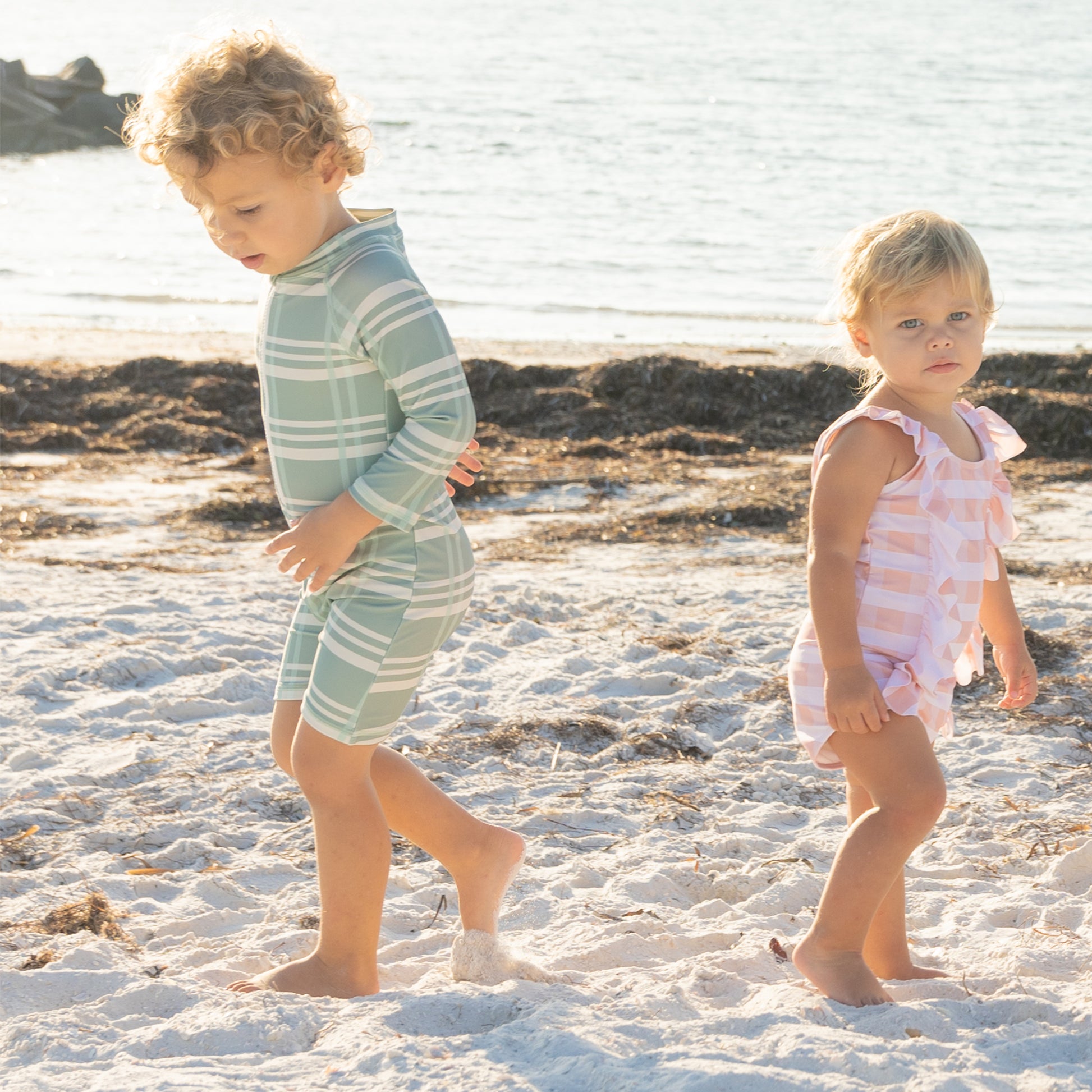 Two children in swimsuits standing on a sandy beach with water in the background