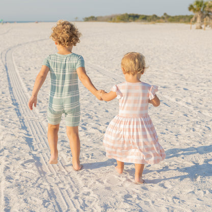 Two young siblings walking together on the beach wearing matching pink and green gingham bamboo outfits.
