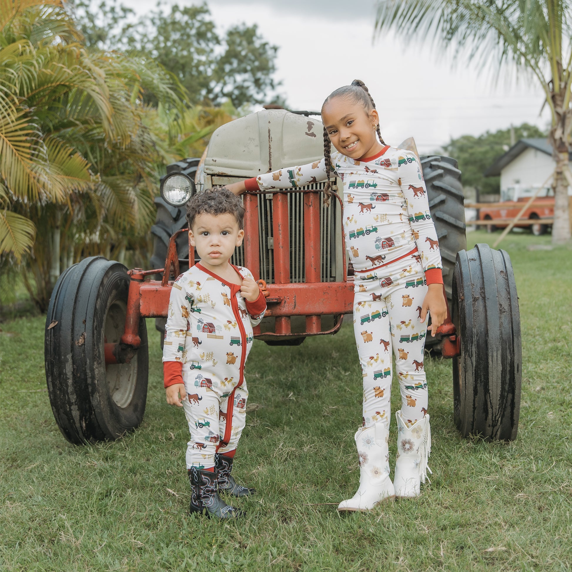 Two kids wearing On the Farm bamboo convertible pajamas with horse, cow, and tractor print, standing near a red tractor.