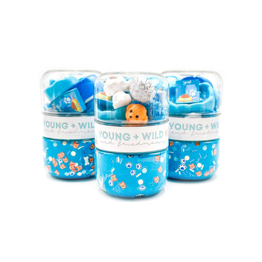 Three clear plastic cylindrical containers of Young + Wild bath and body products arranged in a group on a white background. The containers feature blue lower sections with colorful sea-creature patterns, white bands with the brand name, and colorful contents visible inside, including blue items and a small orange character.