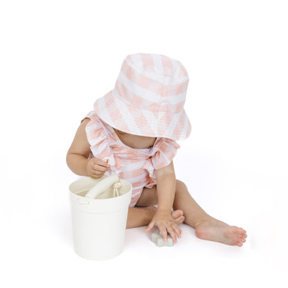 Child wearing a pink and white striped hat and matching outfit, sitting next to a white bucket on a white background.