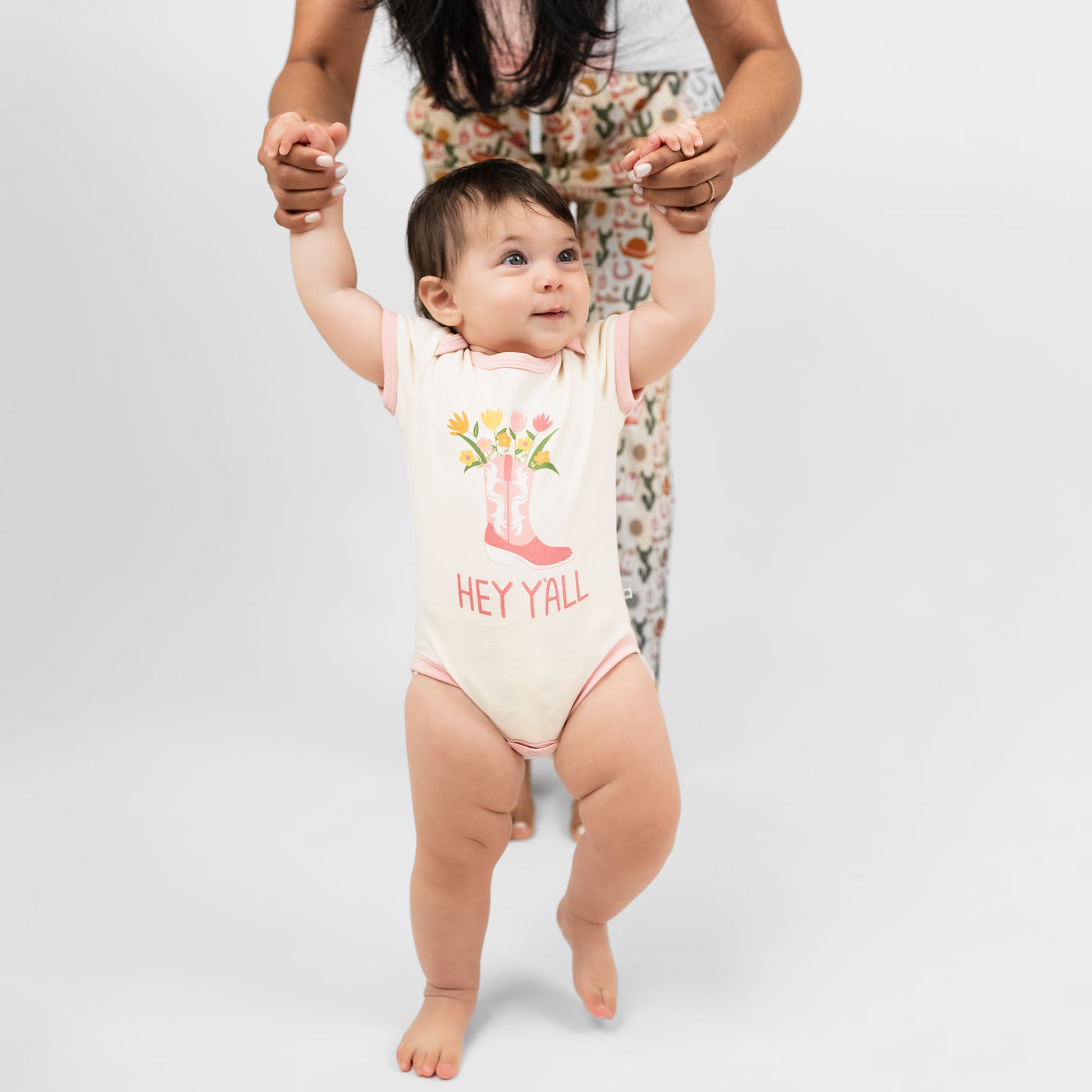 Cute baby girl wearing our Hey Y'all bodysuit paired with a pink cowgirl boots print and floral accents, capturing the essence of rodeo and western style for a perfect outfit.