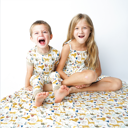 two kids sit on the "jungle friends" bamboo blanket. the boy on the left roars like a lion in his "jungle friends" 2-piece matching pajama set. the girl next to him laughs in her "jungle friends" dress. the "jungle friends" print is a mix of jungle animals that include giraffes, lions, cheetahs, sloths, snakes, and forest plants. 