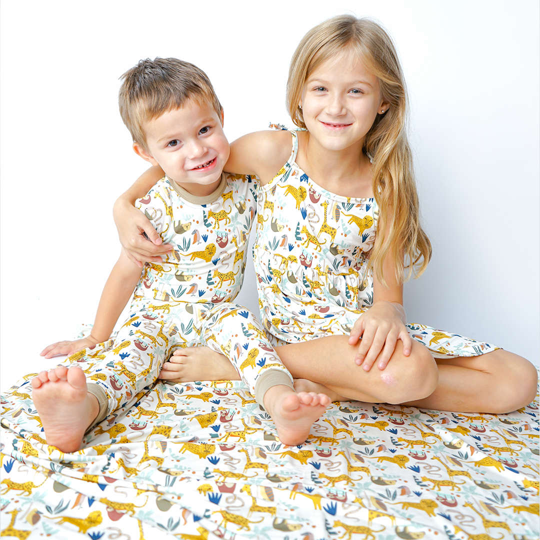 2 kids sit on the "jungle friends" bamboo blanket. she has her arm wrapped around the boy. they are both wearing "jungle friends" apparel. the boy in the matching pajama set and the girl in the dress. the "jungle friends" print is a mix of jungle animals that include giraffes, lions, cheetahs, sloths, snakes, and forest plants. 