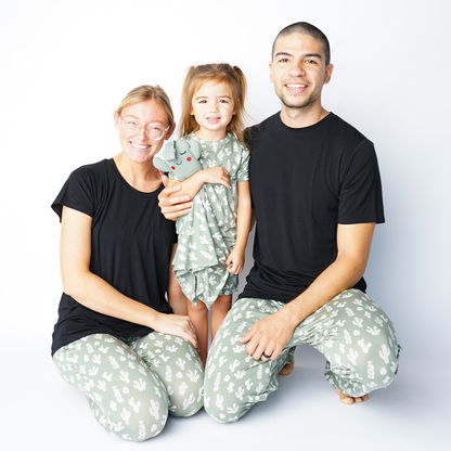 family of 3 smiles in their "stay sharp apparel. Mom and dad sit on the floor and hold on to their baby who stands. mom  is in the jogger pants, dad wears the relaxed pants, and the baby wears the convertible. the "stay sharp" print is a variety of different white cacti on a greyish/green background. 