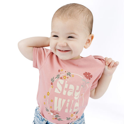 A baby wearing a pink short-sleeve t-shirt with a floral graphic and the words 'Stay Wild' in white, paired with light blue denim jeans, posing against a solid white background.