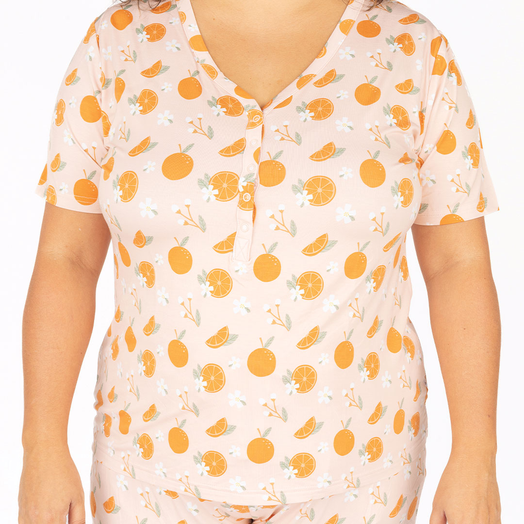 a up close picture of the "freshly squeezed" women's top. the "freshly squeezed" print has an assortment of full and half cut oranges scattered around. there is also flower heads and flower stems that intermingle within the print. this is all space out around a pink background space. 