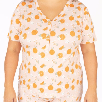 a up close picture of the "freshly squeezed" women's top. the "freshly squeezed" print has an assortment of full and half cut oranges scattered around. there is also flower heads and flower stems that intermingle within the print. this is all space out around a pink background space. 