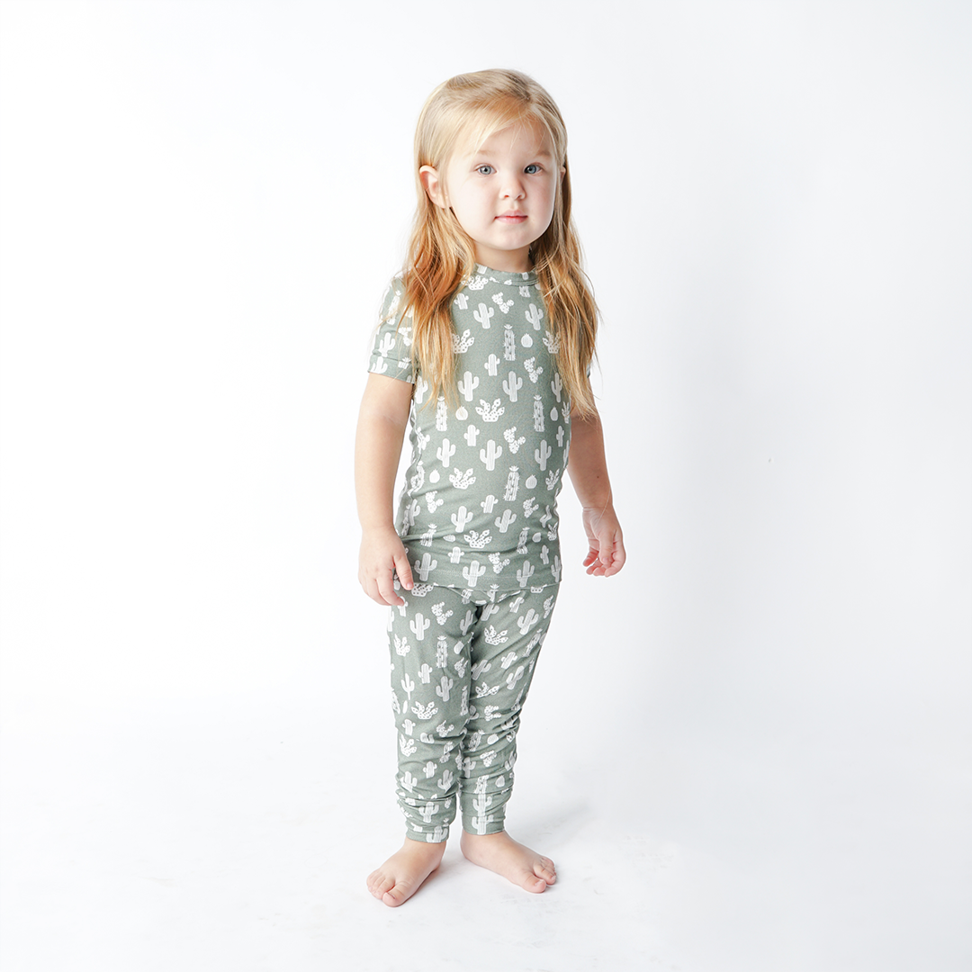 a young girl stands with her arms at her side. she wears the "stay sharp" matching pajama set. the "stay sharp" print is a variety of different white cacti on a greyish/green background. 