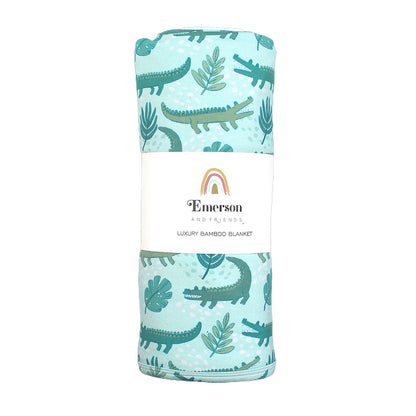 the "later gator" blanket in it's wrapped up packaging. the "later gator" print has a mix of light and dark green alligators, leaves, and white dots scattered on a teal colored background. 