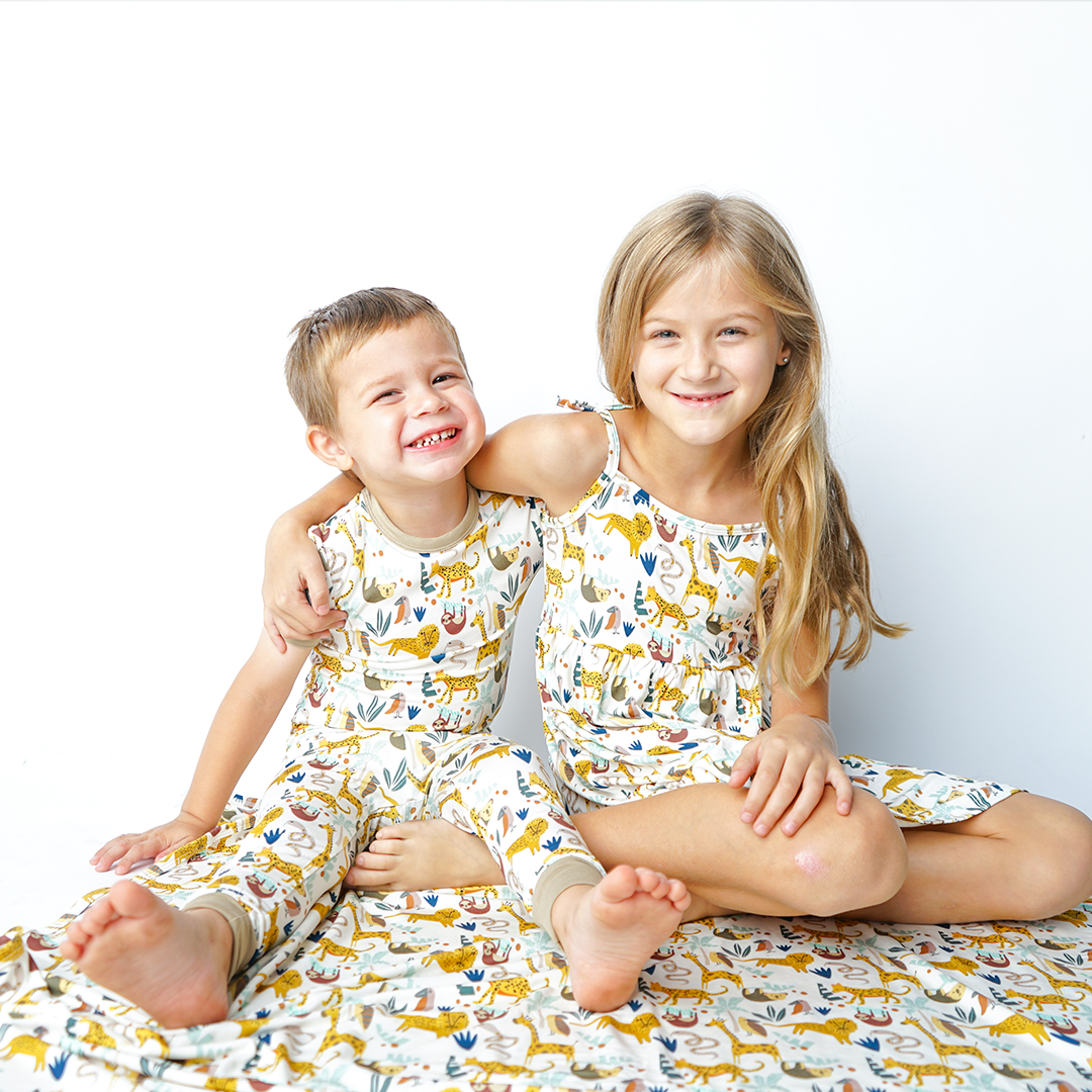 brother and sister sit on the "jungle friends" bamboo blanket. the boy smiles and wraps an arm around his sister. he wears the "jungle friends" matching pajama set. the girl on the right smiles softly and wraps her arm around the brother. she is in the "jungle friends" dress. the "jungle friends" print is a mix of jungle animals that include giraffes, lions, cheetahs, sloths, snakes, and forest plants. 