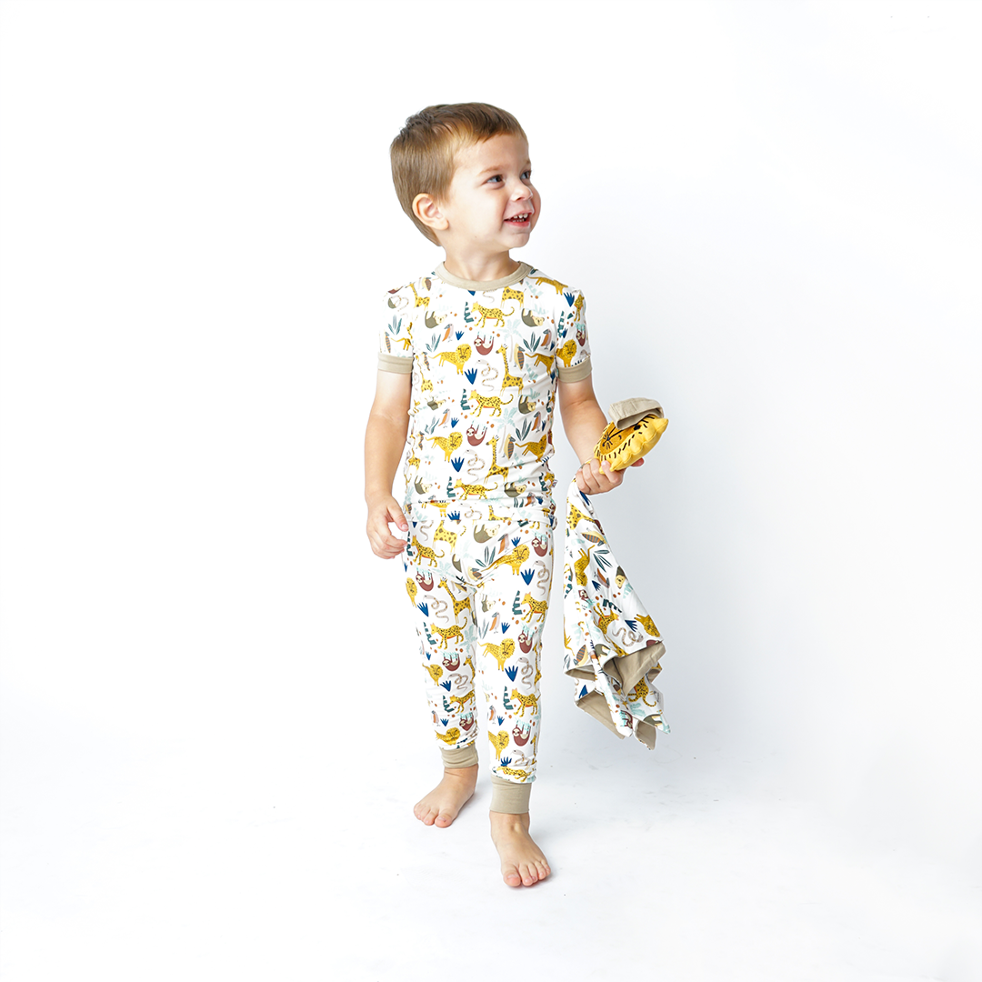 a boy walks and looks at something off camera. he is wearing the "jungle friends" matching pajama set and holds the lion lovey. the "jungle friends" print is a mix of jungle animals that include giraffes, lions, cheetahs, sloths, snakes, and forest plants. 