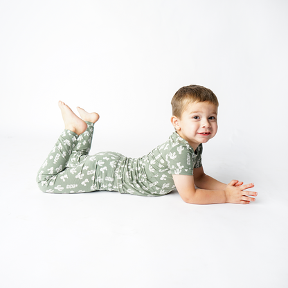 little boy lays on his stomach with his feet in the air. he smiles at the camera. he is wearing the "stay sharp" matching pajama set. the "stay sharp" print is a variety of different white cacti on a greyish/green background. 
