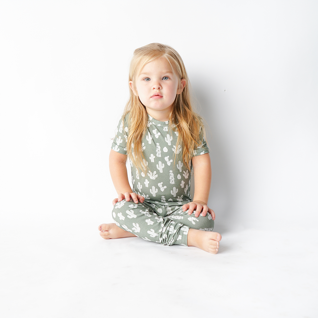 a little girl sits criss-cross on the floor and stares at the camera. she is wearing the 2-piece "stay sharp" matching pajama set. the "stay sharp" print is a variety of different white cacti on a greyish/green background. 