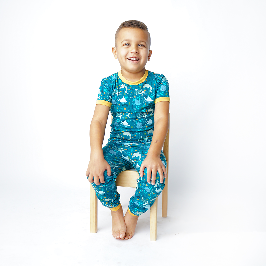 boy sits in a wooden chair wearing his "ocean friends" matching 2-piece set. the "ocean friends" print is a combination of dolphins, stingrays, fish, starfish, coral, bubbles, and sharks, all spread out on a deep sea blue background.