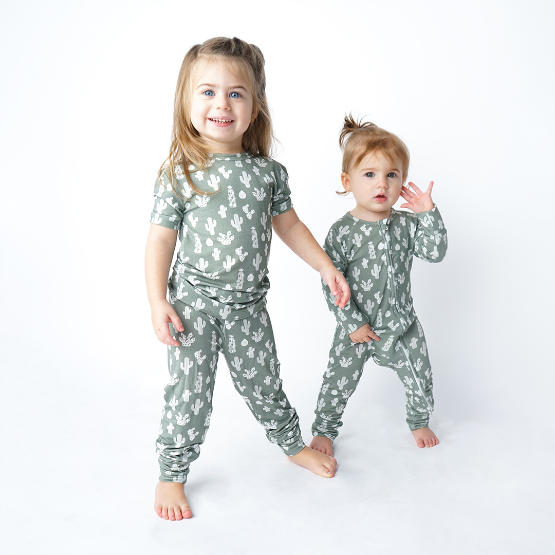 2 girls stand and look at the camera. the older girl is in the "stay sharp" matching pajama set. the younger girl next to her, wears the "stay sharp" convertible. the "stay sharp" print is a variety of different white cacti on a greyish/green background. 